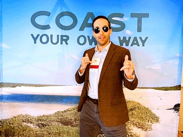 Man in sunglasses posing confidently in front of a backdrop that reads 'Coast Your Own Way'