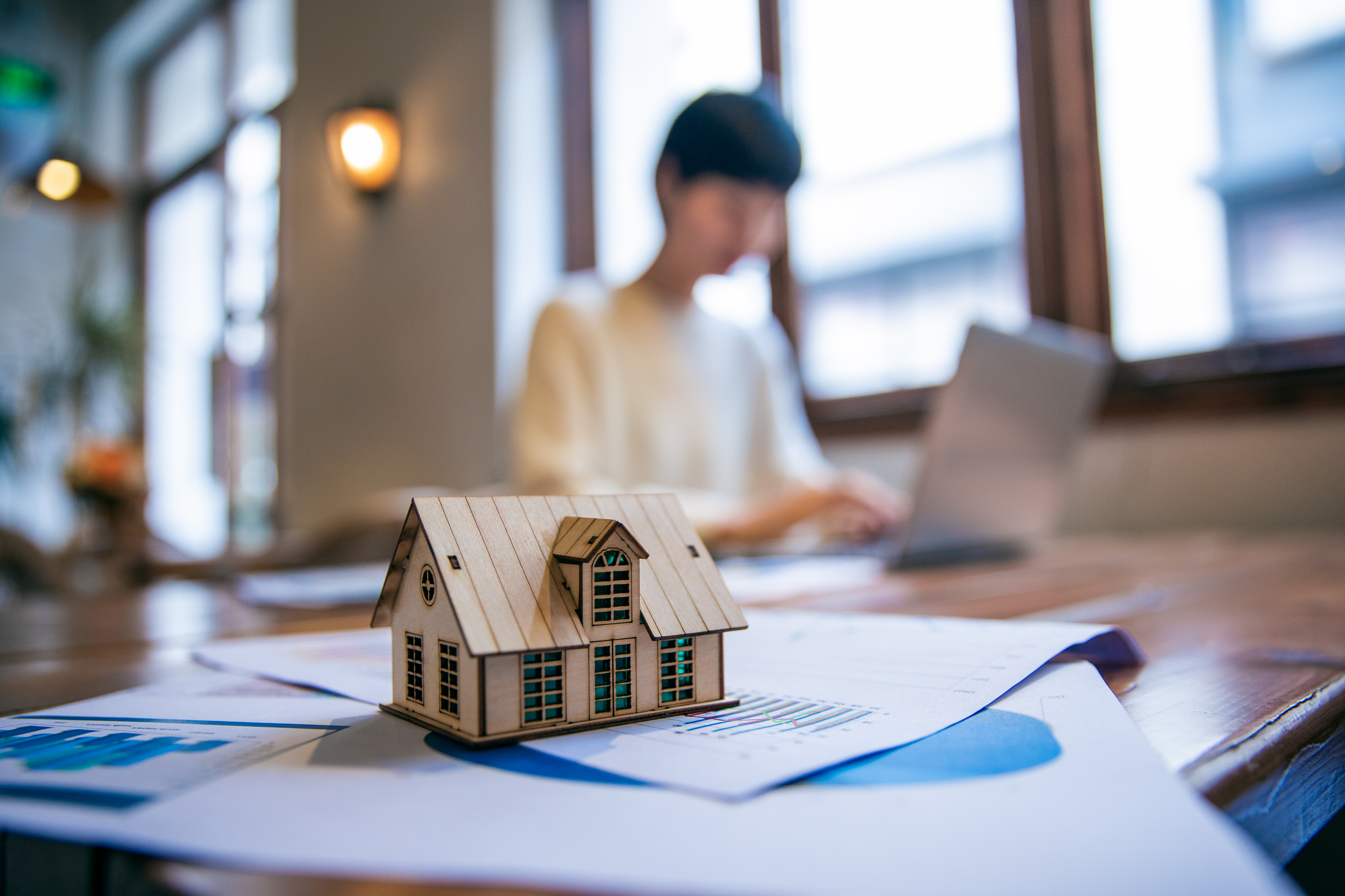Small model house on financial documents with a person working on a laptop in the background